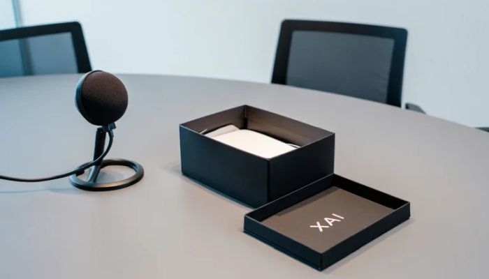 KI-generiertes Symbolbild: Eine offene schwarze Schachtel mit der Beschriftung XAI auf der Innenseite des Deckes liegt auf einem Bürotisch, an dem mit einem Mikrophon und zwei Bürostühlen eine Podcastsituation nachgestellt wird.